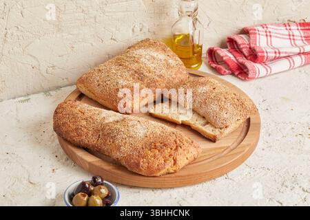 Dinkel-Ciabatta auf Holzoberfläche mit Oliven und Olivenöl im Hintergrund – ein rustikales Brot im mediterranen Stil, perfekt für Aperitif. Stockfoto