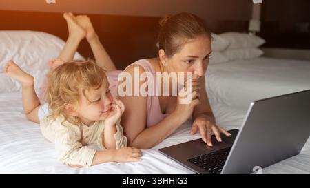 Frau, die an einem Laptop arbeitet und Kind im Hotelzimmer im Bett liegt Stockfoto