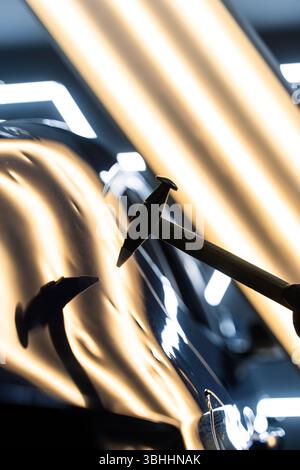 Bei der lacklosen Dellen-Reparatur bei PDR werden Dellen unter Lichtreflexion für Präzision geschlagen. Der Techniker verwendet PDR-Werkzeuge, um Dellen von der Motorhaube zu entfernen Stockfoto