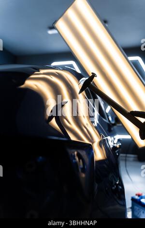Bei der lacklosen Dellen-Reparatur bei PDR werden Dellen unter Lichtreflexion für Präzision geschlagen. Der Techniker verwendet PDR-Werkzeuge, um Dellen von der Motorhaube zu entfernen Stockfoto