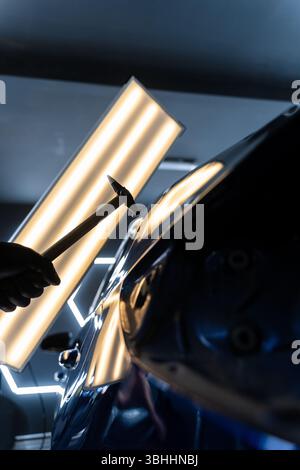 Bei der lacklosen Dellen-Reparatur bei PDR werden Dellen unter Lichtreflexion für Präzision geschlagen. Der Techniker verwendet PDR-Werkzeuge, um Dellen von der Motorhaube zu entfernen Stockfoto