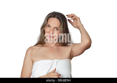 Attraktive lächelnde reife Frau in Handtuch auf weißem Hintergrund gewickelt, die Hand hebt, um Haare zu reparieren, Kopierraum. Körperpflege nach 50 Stockfoto