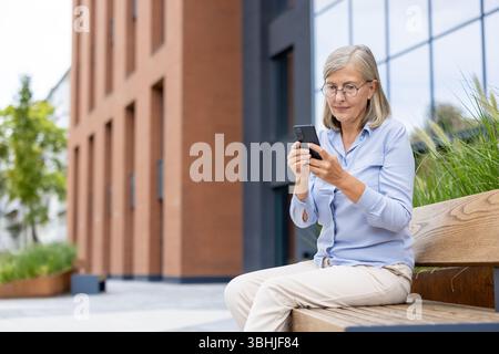 Eine ältere Frau mit einer Brille, die ein Smartphone benutzt, während sie auf einer Bank in einer städtischen Umgebung in der Nähe eines modernen Gebäudes sitzt. Stockfoto