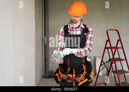 Junger Baumeister in einem Schutzhelm, der Arbeitshandschuhe vor dem Hintergrund einer Wand anzieht. Das Konzept der Bau- und Hausrenovierung Stockfoto