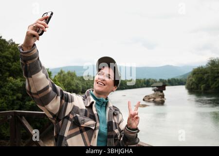 Eine Frau macht ein Selfie mit einem Friedenszeichen mit Drina River und dem Haus auf dem Felsen im Hintergrund. Reisebloggerin. Serbien Land im Frühjahr Stockfoto