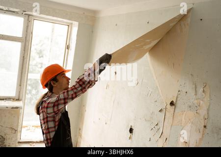 Renovierungsarbeiter in HardHat Entfernen des alten Hintergrundbildes von der Wand während der Wohnungsvorbereitung Stockfoto