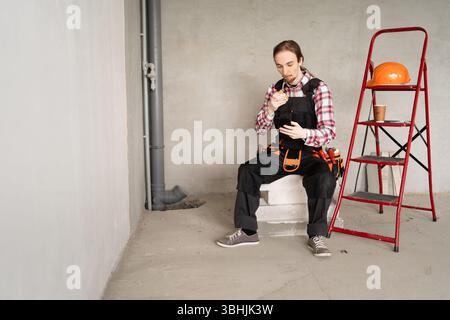 Junger Baumeister isst Burger mit Smartphone drinnen Stockfoto