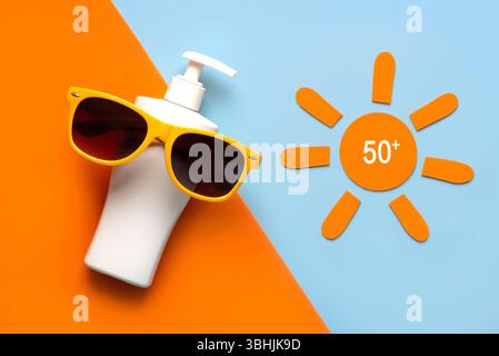 Blick von oben auf eine Flasche Sonnencreme, gelbe Sonnenbrille und orangefarbene Sonne mit 50+ Aufschriften auf orangem und blauem Hintergrund. Konzept des Sonnenschutzes Stockfoto