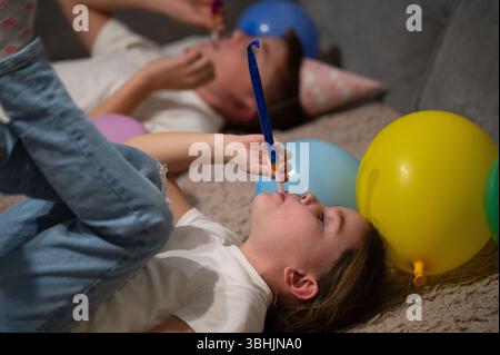 Zwei fröhliche Kinder liegen auf einer weichen Oberfläche, tragen Partyhüte und wehende Hörner, umgeben von lebhaften Ballons, die die Freude eines Celli einfangen Stockfoto