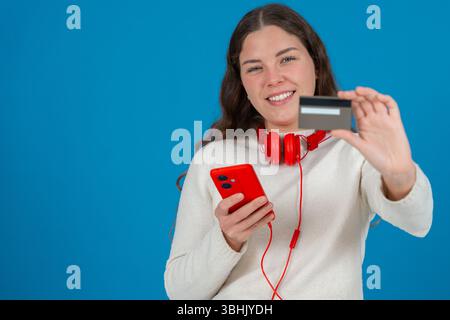 Frau zeigt Kreditkarte vor der Kamera, während sie das Smartphone auf blauem Hintergrund hält Stockfoto