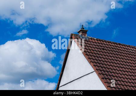Bei einer Nahaufnahme des Dachs des Hauses sind braune Fliesen und eine weiße Außenwand zu sehen. Flauschige weiße Wolken treiben in einem leuchtend blauen Himmel und schaffen eine Ruhe Stockfoto