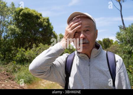 Ein älterer Mann, der unter der Hitze leidet, während er im Wald spaziert und mit der Hand den Schweiß abwischt. Stockfoto