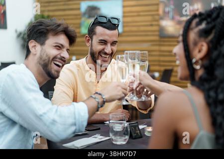 Eine überschwängliche Gruppe verschiedener junger Freunde lacht herzhaft, während sie in einem Restaurant im Freien Gläser Champagner oder Prosecco anrösten und anstoßen. A g Stockfoto