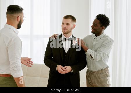 Kaukasischer junger erwachsener Mann im Smoking zwischen schwarzem jungen erwachsenen Mann und kaukasischem jungen erwachsenen Mann, Freunde unterstützen und sprechen während der Hochzeitsfeier drinnen Stockfoto