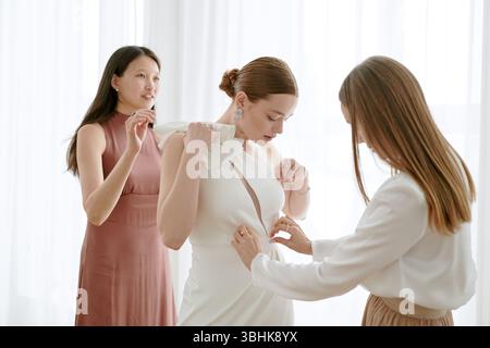 Kaukasische junge Erwachsene Frau steht, während zwei junge Erwachsene Frauen beim Anpassen der Hochzeitskleider helfen, sich auf die Hochzeitsfeier in einem hellen Raum mit transparenten Vorhängen vorzubereiten Stockfoto