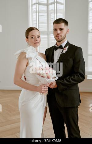 Porträt einer kaukasischen jungen erwachsenen Frau im Hochzeitskleid mit Blumenstrauß neben dem kaukasischen jungen erwachsenen Mann im Anzug, der sie während der Hochzeitsfeier drinnen umschließt Stockfoto