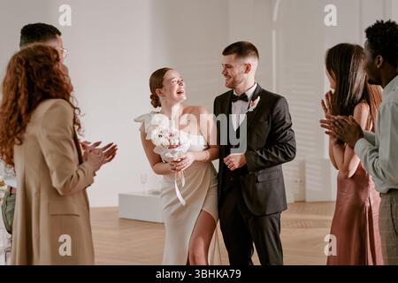 Kaukasische junge erwachsene Braut lacht mit kaukasischem jungen erwachsenen Bräutigam, während er einen Blumenstrauß hält, umgeben von verschiedenen Gruppen junger erwachsener Freunde, die während der Hochzeitsfeier klatschen Stockfoto