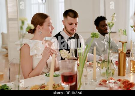 Kaukasische junge Erwachsene Frau sitzt neben kaukasischem jungen erwachsenen Mann und schwarzem jungen erwachsenen Mann am Hochzeitsfeierungstisch, führt Gespräche und genießt gemeinsam ein festliches Essen Stockfoto