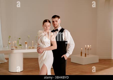Kaukasische junge Erwachsene Frau in elegantem Kleid, die neben kaukasischem jungen erwachsenen Mann in formellem Anzug steht und sich während der Hochzeitsfeier im dekorierten Innenraum posiert Stockfoto