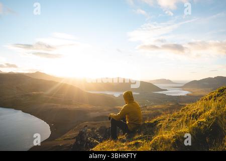 Der Reisende in einer gelben Jacke sitzt auf dem Gipfel des Horns auf der Halbinsel Snaefellsnes in Island und beobachtet den Sonnenuntergang über vulkanischen Tälern und Friedhöfen Stockfoto