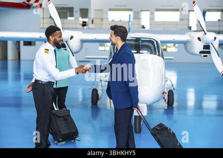 Afrikanischer Pilot schüttelt sich die Hand mit einem Geschäftsmann in einem Flugzeughangar, der beide Trolley-Taschen hält, bereit für einen Privatflug Stockfoto