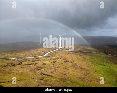 Drohnenaufnahme von Windturbinen unter einem pulsierenden Regenbogen mit näherem Regen – Natur und erneuerbare Energie in dynamischer Harmonie. Stockfoto