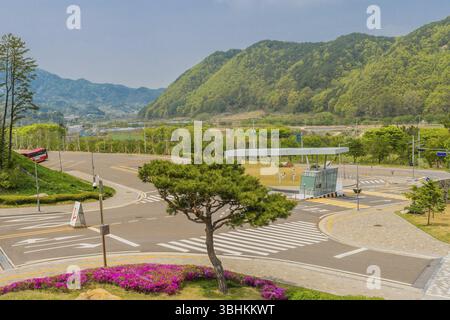 Muju, Südkorea. 1. Mai 2019: Getrimmter immergrüner Baum und violette Azaleen an der Ecke des Eingangstors zum Taekwondowon mit Bergen im Hintergrund Stockfoto