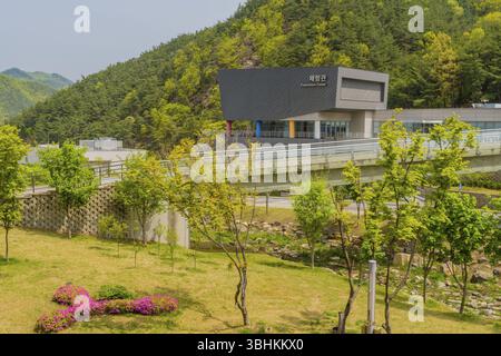 Muju, Südkorea. 1. Mai 2019: Landschaft mit Rasen und trockenem Flussbett, das unter einer Brücke verläuft, mit dem Taewondo Experience Center vor den üppigen Bergen Stockfoto