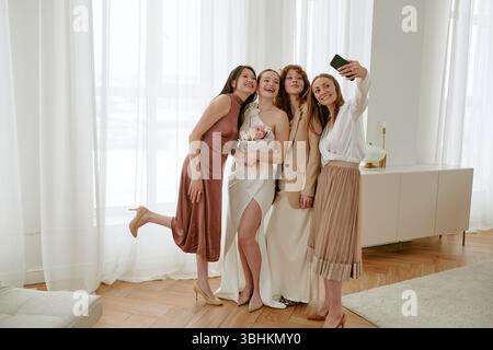 Gruppe von vier jungen kaukasischen Frauen, die zusammen für ein Selfie während der Hochzeitsfeier posieren, Braut mit Blumenstrauß, Freunde lächeln und in hellem Raum stehen Stockfoto