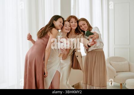 Drei junge Erwachsene kaukasische Frauen und eine junge Erwachsene asiatische Frau lächeln und posieren zusammen für Selfie während der Hochzeitsfeier, Braut mit Blumenstrauß, in der Nähe drinnen Stockfoto