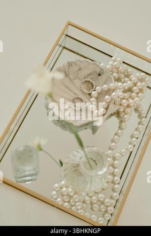 Großansicht mit Eheringen, die in einer dekorativen Schüssel neben einer Perlenschnur und einer Glasvase mit Blume auf einem Spiegeltablett liegen und elegante Details für die Hochzeitsfeier festhalten Stockfoto