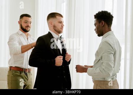 Kaukasischer junger erwachsener Mann, der im Anzug steht und von Schwarzen jungen erwachsenen Männern und jungen erwachsenen Männern unterstützt wird, die sich auf die Hochzeitsfeier im Haus vorbereiten Stockfoto