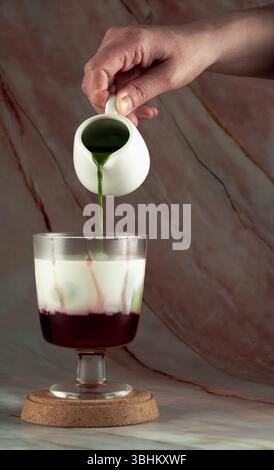 Eine Hand gießt grünen Matcha in ein Glas, das eine rote Schicht Erdbeere enthält, um ein optisch ansprechendes, mehrschichtiges Getränk zu kreieren Stockfoto