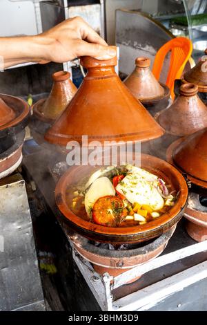 Agadir, Souss-Massa - Marokko - 07-31-2024: Heben Sie den Deckel von einem Taxine-Topf mit der Hand ab, um darin dampfendes Gemüse zu enthüllen. Stockfoto