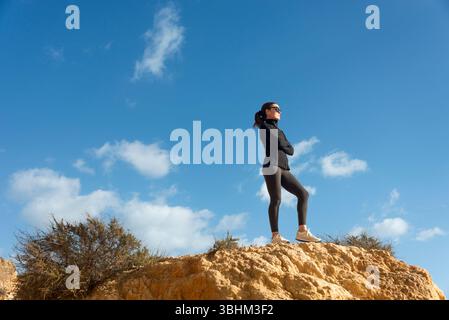 Sportliche, selbstbewusste Frau, die auf Rocky Cliff unter einem hellen blauen Himmel steht Stockfoto