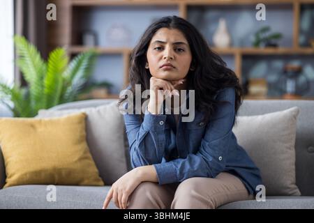 Eine Frau schaut direkt in die Kamera, während sie auf einer Couch sitzt und mit Händen ihr Kinn stützt. Sie hat einen ernsten, nachdenklichen Ausdruck. Stockfoto