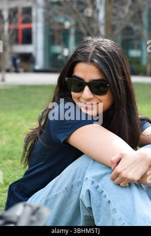 Porträt einer jungen glücklichen Frau, die auf grünem Gras im Stadtpark lächelnd sitzt Stockfoto
