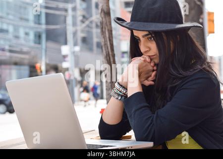 Junge Frau mit Fedorahut, die im Straßencafé sitzt und einen Laptop benutzt Stockfoto
