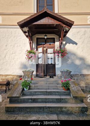 Rustikale alte Gebäudefassade mit Veranda und einer charmanten Holztür, eingerahmt von zwei Seitenfenstern und einem kleinen Überhang. Steintreppen führen zum Eingang Stockfoto