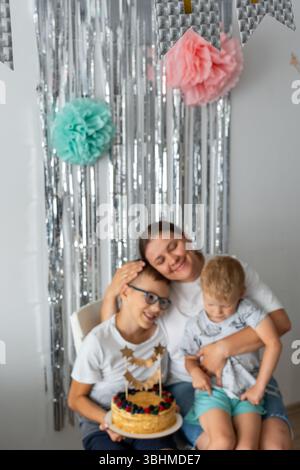 Liebevolle Mutter mit zwei Söhnen, die hausgemachte Geburtstagskuchen mit frischer Obstdekoration und glitzerndem Hintergrund genießen. Ein warmer Familienmoment zu Hause Stockfoto