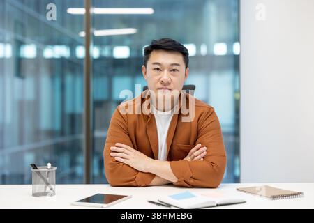 Ein asiatischer Mann in braunem Hemd und weißem T-Shirt, der mit überkreuzten Armen an einem Schreibtisch sitzt. Stockfoto