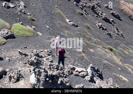 Linguaglossa, Sizilien - Italien - 05-01-2025: Zwei Wanderer gehen bergauf auf auf einem schwarzen vulkanischen Hang mit verstreuten Felsen und kleinen grünen Moosflecken Stockfoto