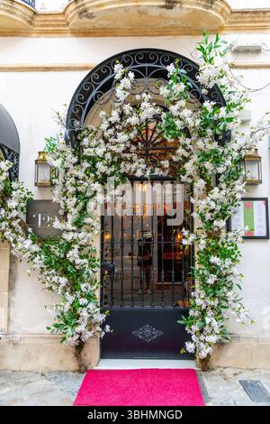 Jerez de la Frontera, Cadiz - Spanien - 23.04.2024: Eingang eines eleganten Gebäudes mit kunstvollen Toren, umgeben von weißen Blumen und einem roten Auto Stockfoto