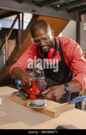 Afroamerikanischer Mann mit Schwingschleifer auf Bank in Holzwerkstatt mit Schutzbrille und Gehörschutz Stockfoto
