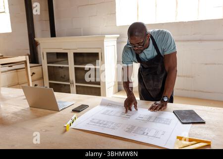 Afroamerikaner, der eine Schürze trägt, beugt sich über die Baupläne auf der Werkbank, indem er Ebenen, Kopierraum benutzt Stockfoto