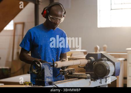 Afroamerikanischer Mann mit Gehörschutz und Schutzbrille, der Holz mit Meißel auf Drehmaschine in der Werkstatt rasiert Stockfoto