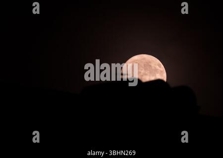 Stuttgart, Deutschland. Juni 2025. Der Mond ist hinter einem Hausdach zu sehen. In der Nacht vom 10. Auf den 11. Juni taucht ein seltenes Himmelsphänomen auf: Der Erdbeermond. Der Vollmond ist dann ungewöhnlich tief am Himmel - so tief wie vor 18 Jahren. Quelle: Markus Lenhardt/dpa/Alamy Live News Stockfoto