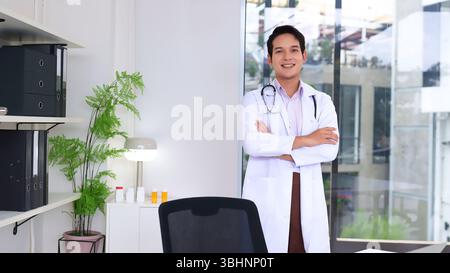 Ein zuversichtlicher, erwachsener asiatischer Arzt steht in ihrem Büro. Sie lächeln, tragen einen weißen Mantel und ein Stethoskop. Es ist ein professionelles Umfeld für Sanitäter Stockfoto