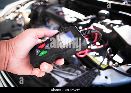 Diagnosewerkzeug für Fahrzeugbatterien: Ein Techniker hält einen Fahrzeugbatterietester über einer geöffneten Motorhaube, wobei der Motor sichtbar ist, um zu prüfen, ob die Fahrzeugbatterie vorhanden ist Stockfoto
