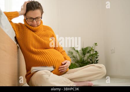 Heitere schwangere Frau schreibt wichtige Liefererinnerungen zu Hause im Notizbuch. Durchdachte Vorbereitung auf die Grundlagen der Entbindungs- und Neugeborenenpflege. Stockfoto